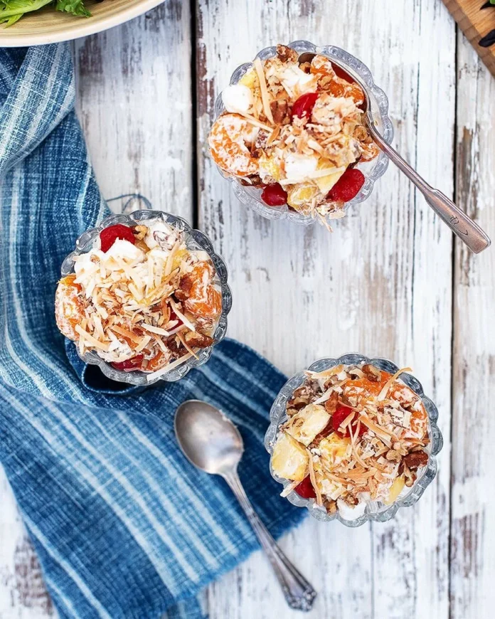 Three small cups of Ambrosia Salad with cherries on top sit on a white picnic table with a blue towel on it.