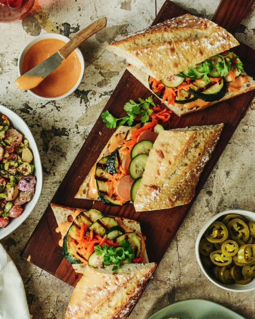 Three baguette sandwiches with grilled zucchini and other veggies sit on a wood cutting board beside sauces and toppings.