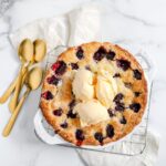A Blackberry Cobbler with three scoops of vanilla ice cream in a white dish sit beside three golden spoons on a white marble counter.