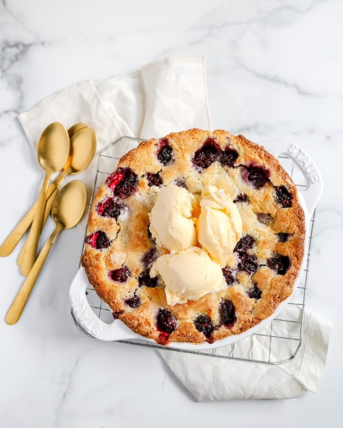 A Blackberry Cobbler with three scoops of vanilla ice cream in a white dish sit beside three golden spoons on a white marble counter.