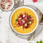 An aerial shot of an orange soup in a white bowl, topped with cucumber and tomato slices and a few colorful sprouts. Cantaloupe Peach Gazpacho