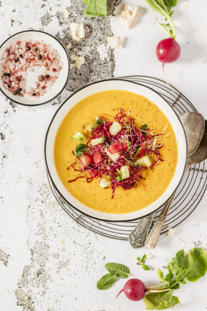 An aerial shot of an orange soup in a white bowl, topped with cucumber and tomato slices and a few colorful sprouts. Cantaloupe Peach Gazpacho