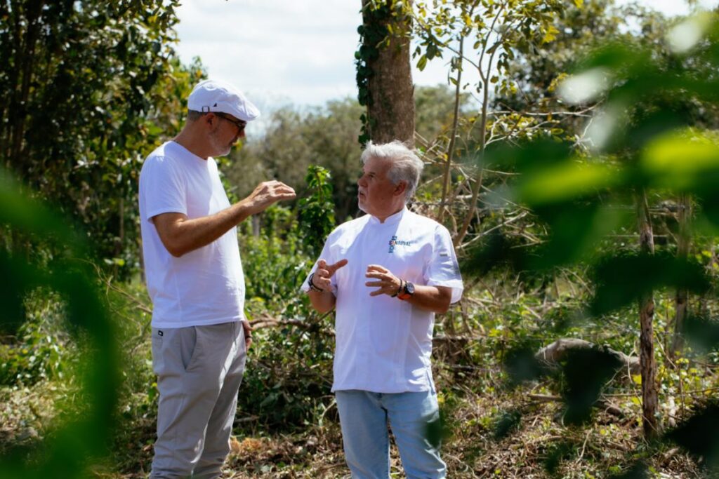 Two men, both wearing white shirts, stand in a green, leafy outdoor area, engaged in conversation.