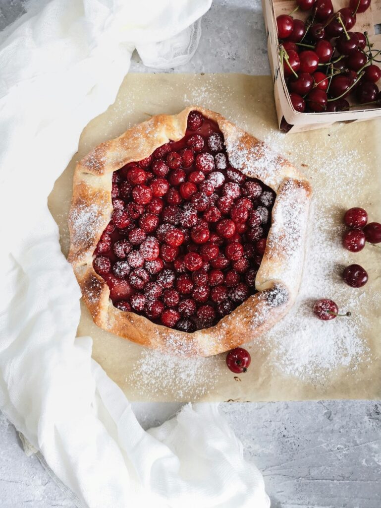A Cherry Galette sits open faced on a countertop with powdered sugar and cherries everywhere.