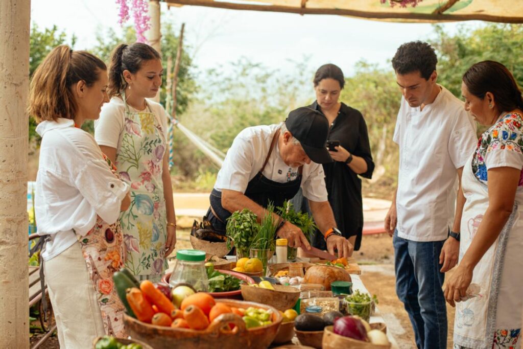 A group of six people, including a chef in a black cap and apron, are gathered around a table outdoors, observing as the chef prepares food.