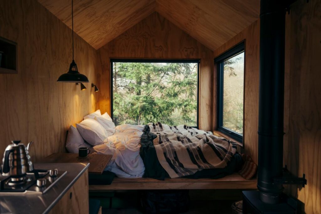 The interior of a cozy wooden cabin features a bed with white and striped bedding, large windows looking out onto a dense forest