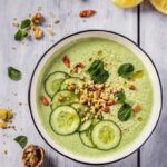 A bowl of green cucumber and yogurt soup with garnishes with seasonings and lemons surrounding the bowl.