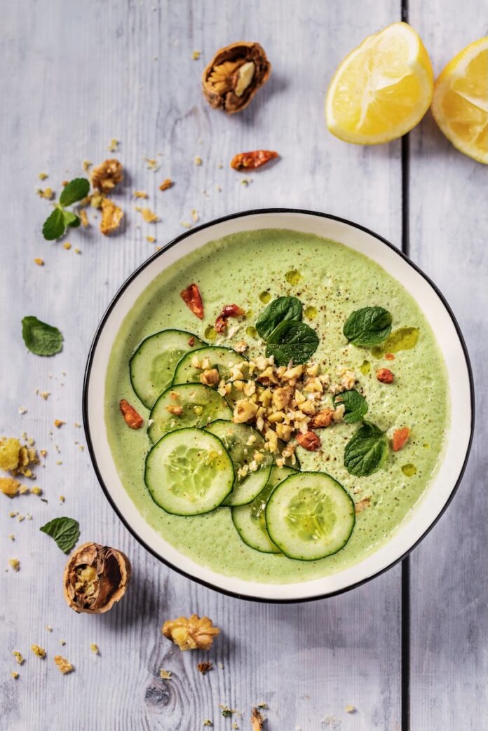 A bowl of green cucumber and yogurt soup with garnishes with seasonings and lemons surrounding the bowl.