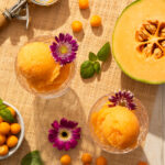 An aerial view of Husk Cherry and Melon Sorbet, an orange ice cream, scooped into two glass containers. Half a melon sits off to the side.