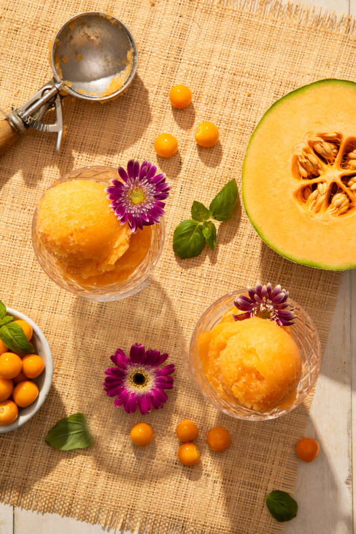 An aerial view of Husk Cherry and Melon Sorbet, an orange ice cream, scooped into two glass containers. Half a melon sits off to the side.