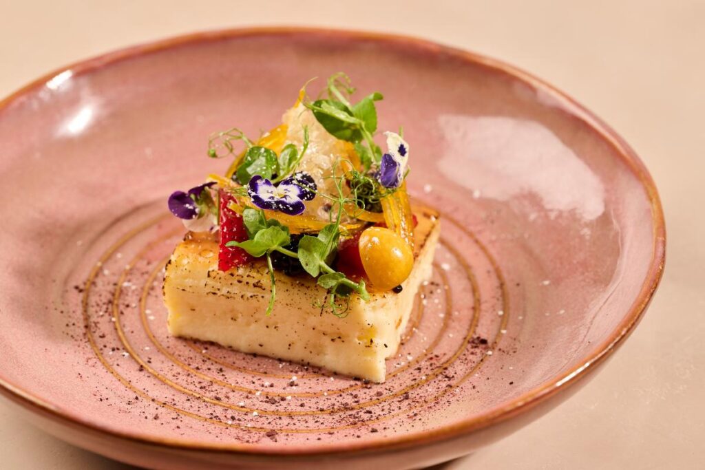 A honey cheesecake, garnished with berries, edible flowers, and candied orange peel, is presented on a plate.