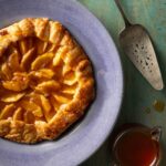 A round Easy Hot Honey Peach Galette sits on a blue plate with a pie spatula sitting beside it.