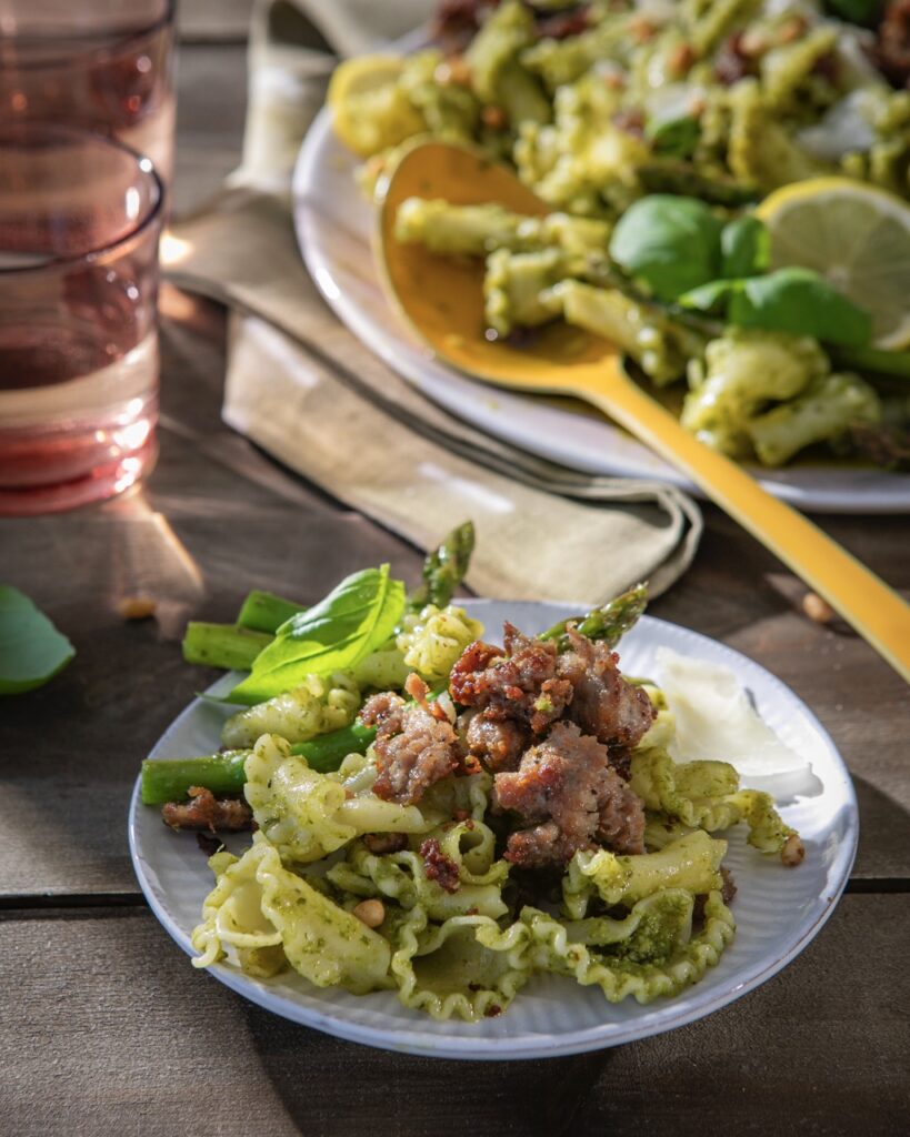 A small plate of Easy Pesto Pasta with basil, sausage, pesto sauce, and asparagus as a large plate of the dish sits in the background.