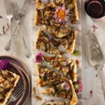 An eggplant and smoked mozzarella tart cut into pieces on a white table beside silverware and plates.