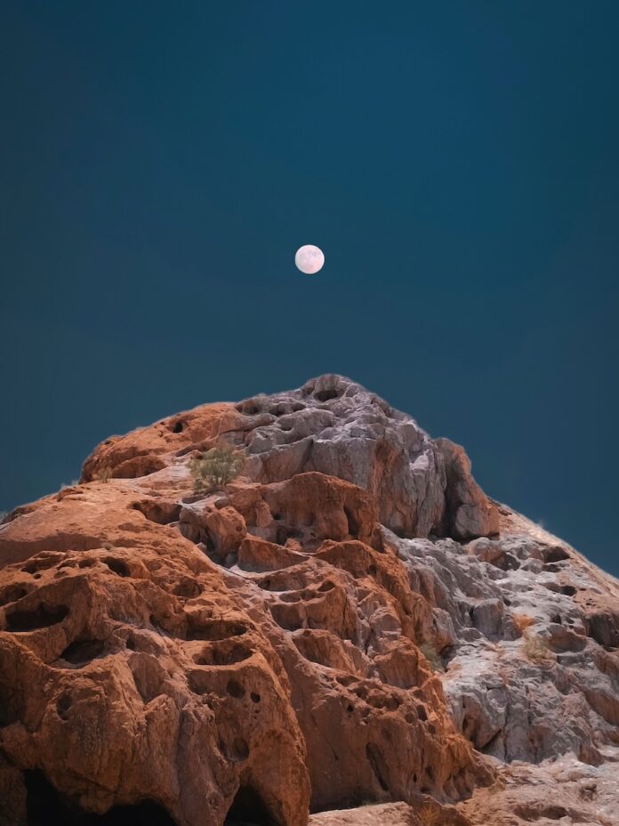 A full moon in July over an orange, rocky mountain.