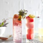 Two craft mocktails sit side by side, one with watermelon cubes, the other with grapefruit and herbs.