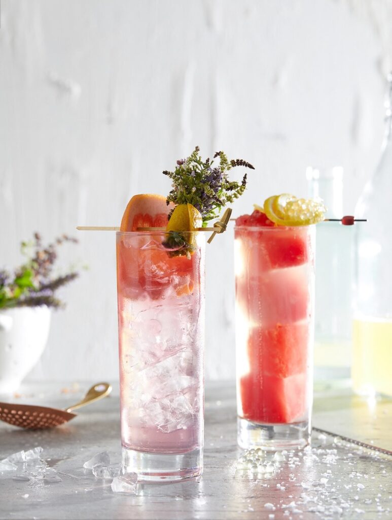 Two craft mocktails sit side by side, one with watermelon cubes, the other with grapefruit and herbs.