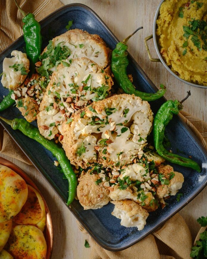 Slices of Gobi Musallam, spiced whole cauliflower, sit on a baking sheet with whole green chiles.