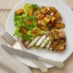 A white plate with a piece of grilled swordfish with a pineapple salsa on top and a salad on the side. A fork and knife also sit on the plate.