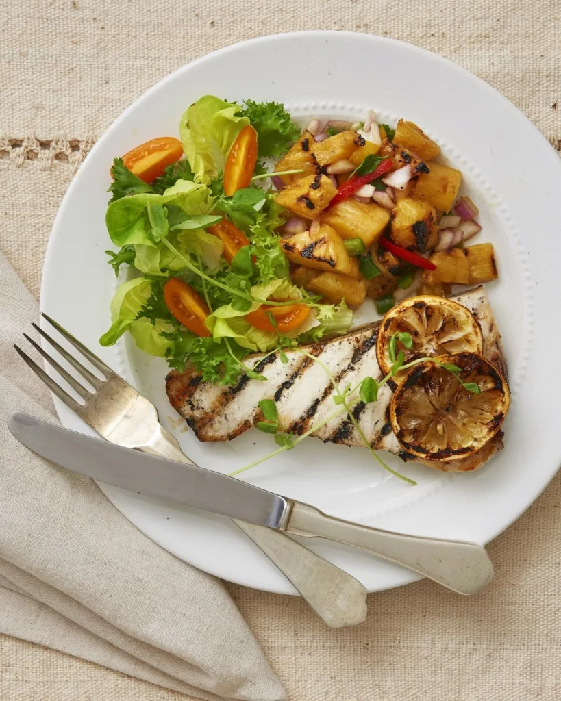 A white plate with a piece of grilled swordfish with a pineapple salsa on top and a salad on the side. A fork and knife also sit on the plate.