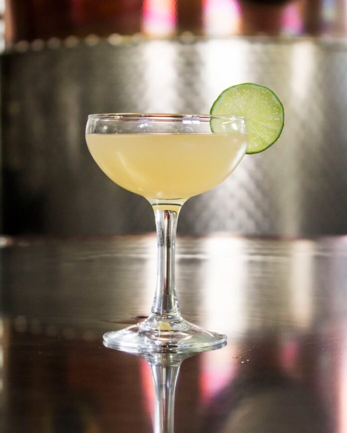 A cocktail glass with a Hemingway Daiquiri featuring a lime wheel garnish on a metal counter.
