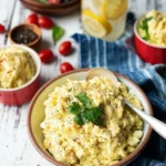 A bowl of Hillbilly Potato Salad beside two smaller bowls with a parsley garnish on top, all on a picnic table.