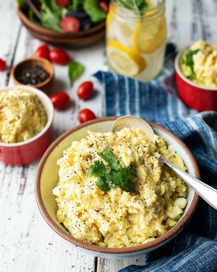 A bowl of Hillbilly Potato Salad beside two smaller bowls with a parsley garnish on top, all on a picnic table.