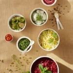 Various bowls of homemade burger toppings sit in white containers on a brown table surrounded by herbs and spices on the table.