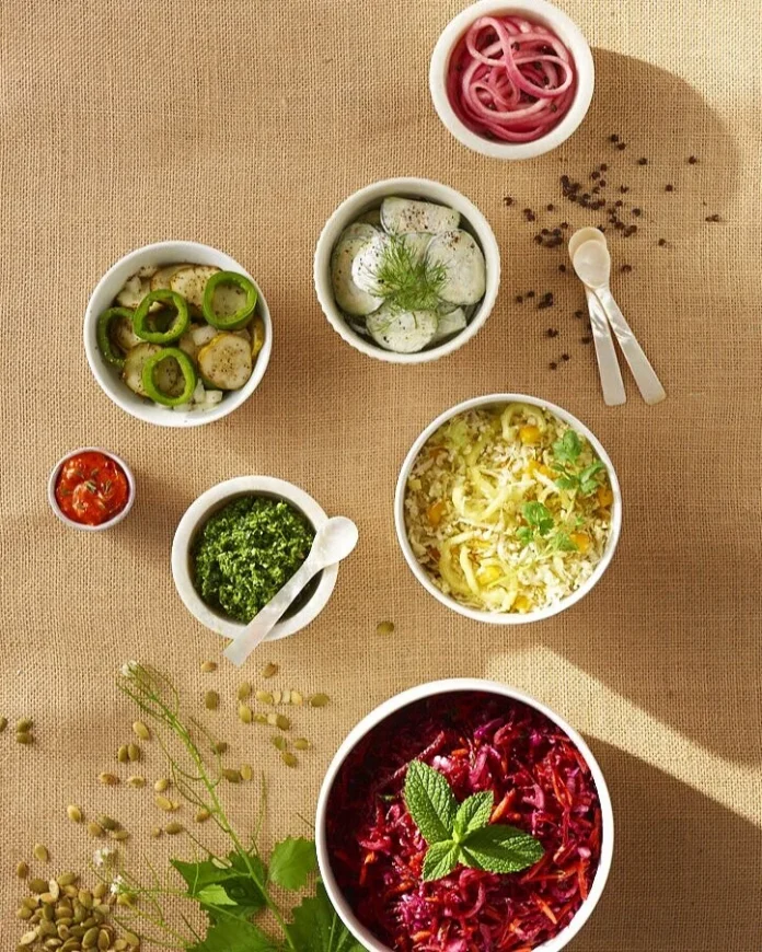Various bowls of homemade burger toppings sit in white containers on a brown table surrounded by herbs and spices on the table.