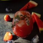 A short glass of homemade watermelon juice with ice and sprigs of lavender on top as fruit ice cubes and watermelon slices sit on the table.