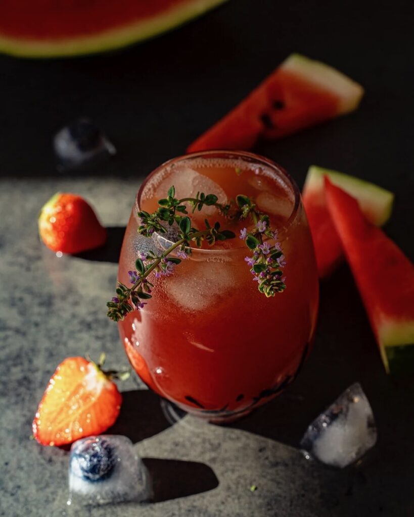 A short glass of homemade watermelon juice with ice and sprigs of lavender on top as fruit ice cubes and watermelon slices sit on the table.