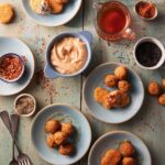 Various small plates of appetizers like chicken nuggets and falaffel surround a small bowl of Hot Honey Aioli with Harissa.