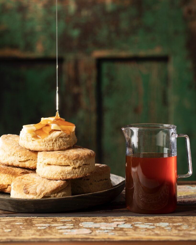 A drizzle of hot honey gets poured onto a stack of homemade buttermilk biscuits sitting beside a pitcher of hot honey.