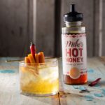 A short glass holds a Hot Honey Old Fashioned with an orange slice and chili pepper garnish as a bottle of Mike's Hot Honey sits to the right of it.