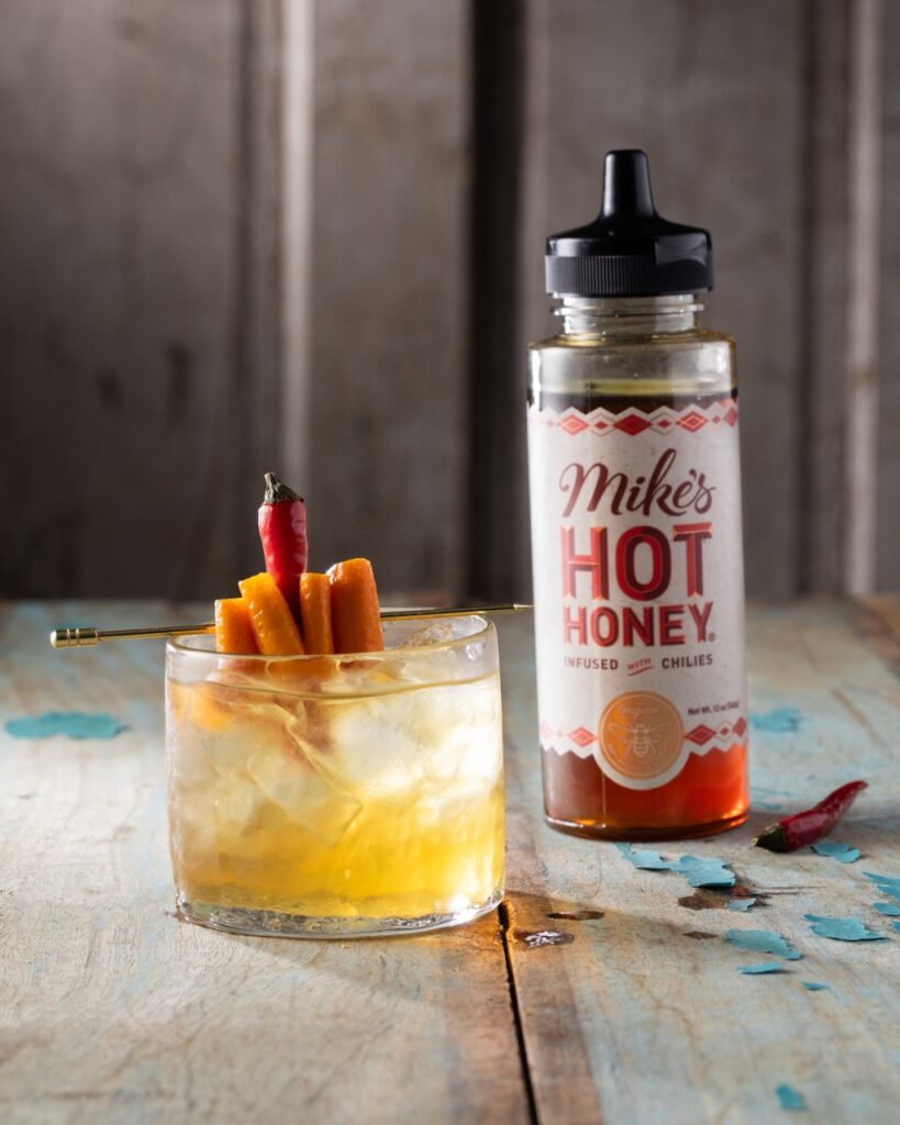 A short glass holds a Hot Honey Old Fashioned with an orange slice and chili pepper garnish as a bottle of Mike's Hot Honey sits to the right of it.