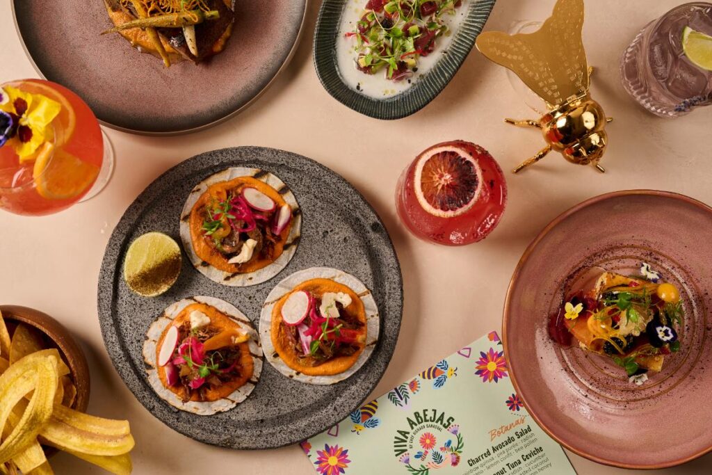 A table is laden with various dishes, including tacos, a salad with edible flowers, and colorful cocktails.