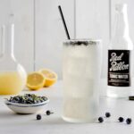 A juniper lemonade tonic sits on a table with juniper berries for a garnish and around it as well as lemons and a bottle of red ribbon soda.