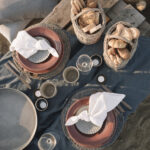 A picnic table holds four wine glasses, two baskets of bread, and two orange place settings.