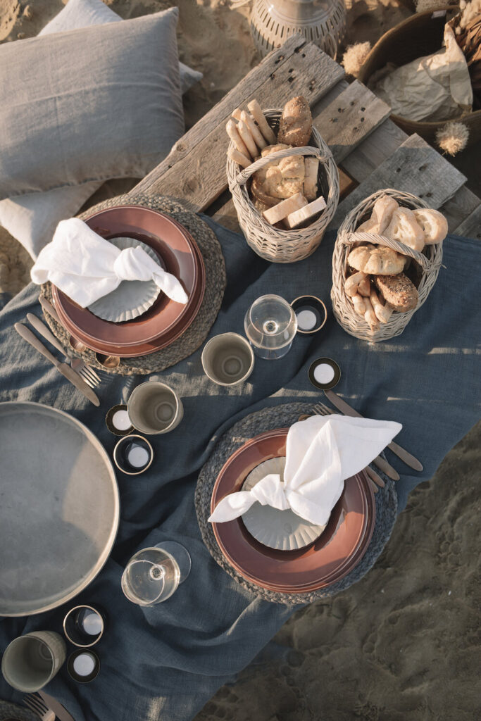 A picnic table holds four wine glasses, two baskets of bread, and two orange place settings.