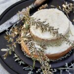 A white buttercream naked cake with two layers sits on a black tray beside a knife and with herbs draped over it.