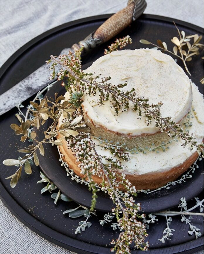 A white buttercream naked cake with two layers sits on a black tray beside a knife and with herbs draped over it.