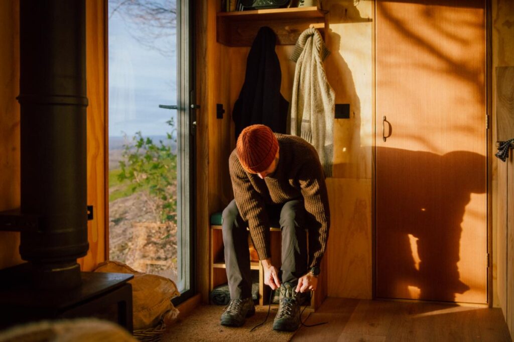 A man wearing a brown knitted sweater and an orange beanie sits on a wooden bench, tying the laces of his hiking boots, with a large window looking out onto a landscape behind him.
