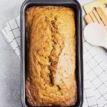 A loaf of Pawpaw Sweet Bread in an orange color sits in a loaf pan on a white towel and counter.