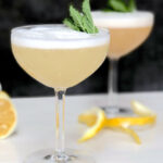 Two tall glasses hold a Pisco Sour with a mint leaf garnish and orange peels on the counter.
