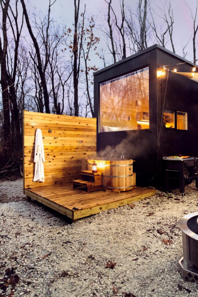 A modern black cabin with a large window is situated on a wooden deck in a forest.