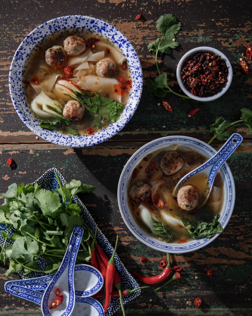 A large blue and white bowl of quick and easy wonton soup sits diagonal to a smaller bowl that has a wonton soup spoon inside it. Both bowls are surrounded by greens and chili peppers.