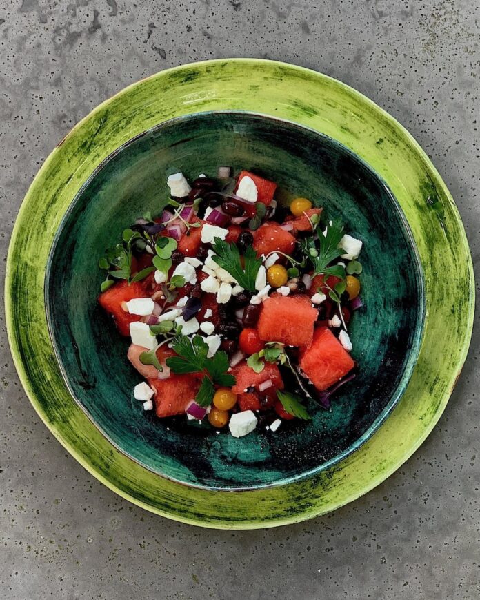 A green plate holds a rose watermelon salad with microgreens, beans, onions, tomatoes, and watermelon.