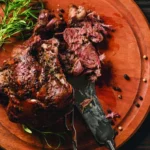A leg of lamb with a dry rub santa maria-style seasoning sits on an orange plate with a fork and knife cutting it open.