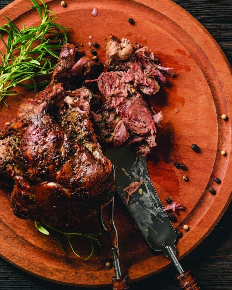 A leg of lamb with a dry rub santa maria-style seasoning sits on an orange plate with a fork and knife cutting it open.