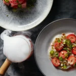 To the right side of the photo sits Scallop Carpaccio dressed in Chilled Strawberry Vinaigrette on a white plate.